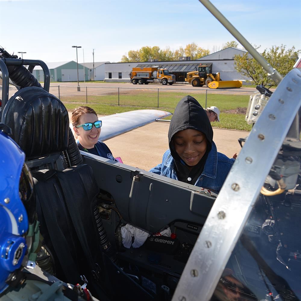  student looking inside a cockpit with smiling teacher in the background